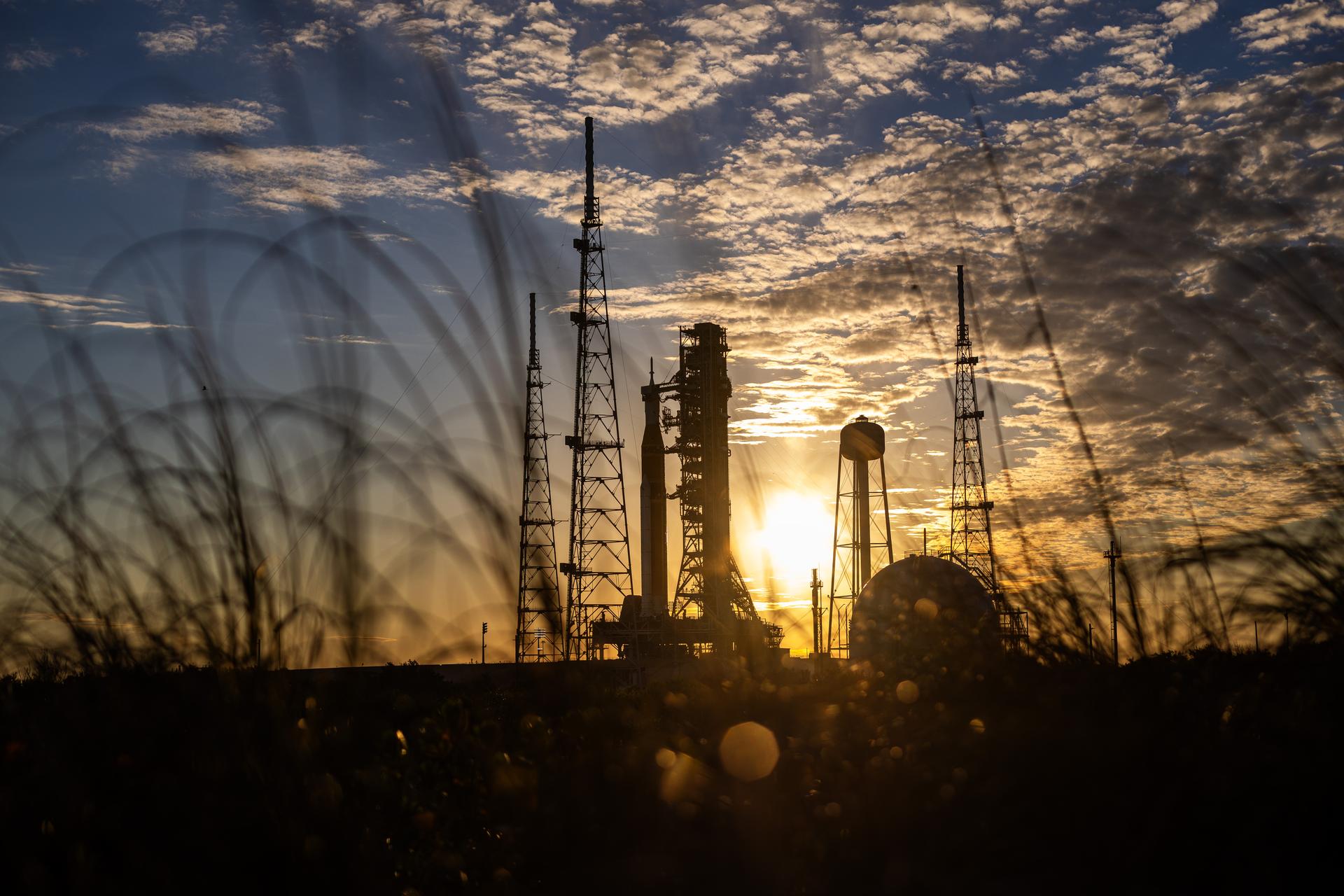 The sun sets behind NASA’s Space Launch System (SLS) rocket and Orion spacecraft as they stand fully assembled atop the mobile launcher at Launch Pad 39B at NASA’s Kennedy Space Center in Florida. The sky glows with warm shades of orange and pink, silhouetting the towering rocket and its solid rocket boosters against the fading light. Photographed on January 31, 2026, the scene captures teams preparing for a wet dress rehearsal for the Artemis II mission, rehearsing launch countdown timelines and procedures as day turns to night.